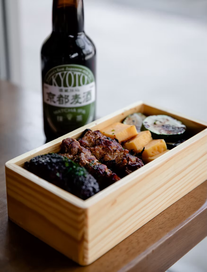 Wooden bento box of skewers and vegetables beside a bottle of Kyoto craft beer