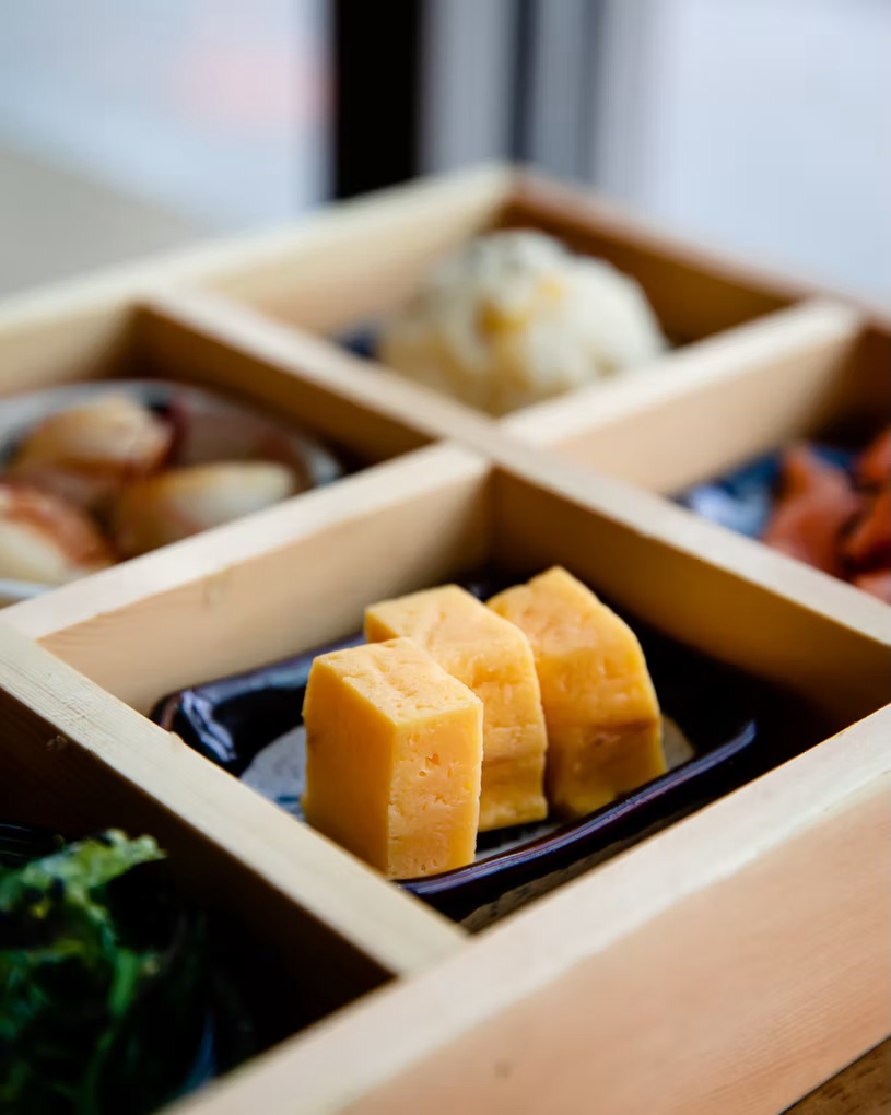 Bento compartments with tamagoyaki, scallops and greens in a wooden box