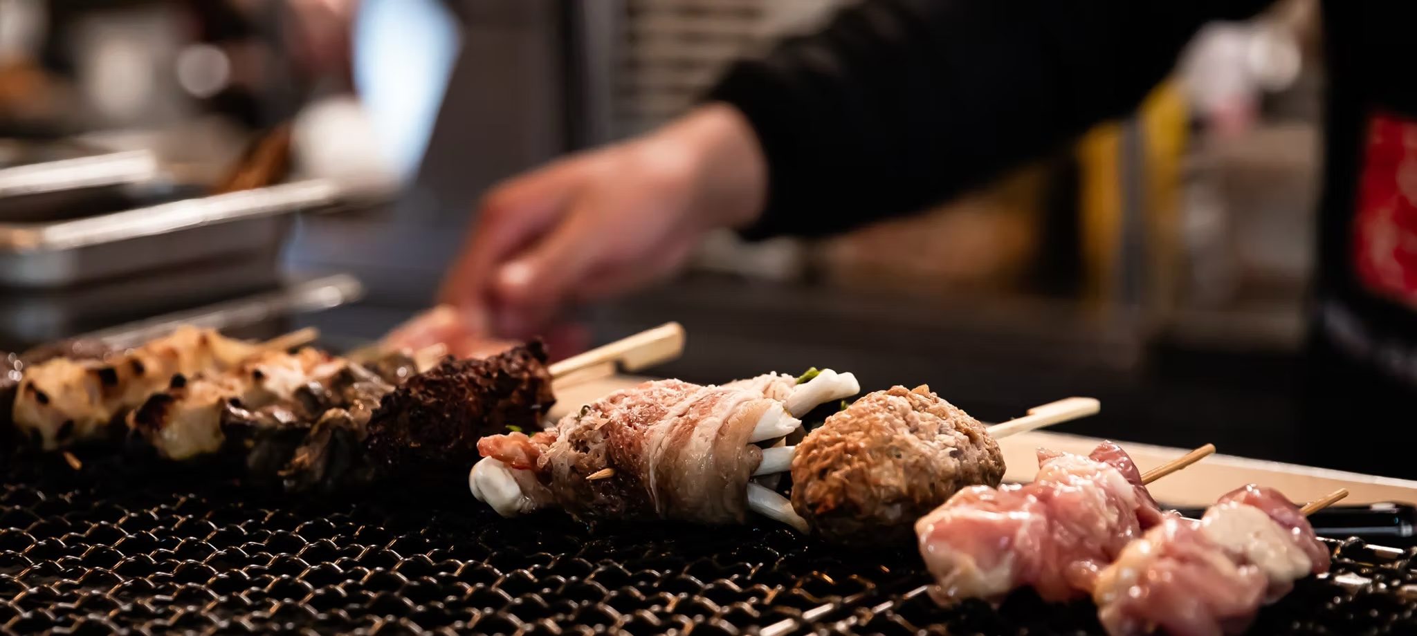 Chef's hand turning skewers over the charcoal grill, bacon-wrapped rolls and chicken lined up