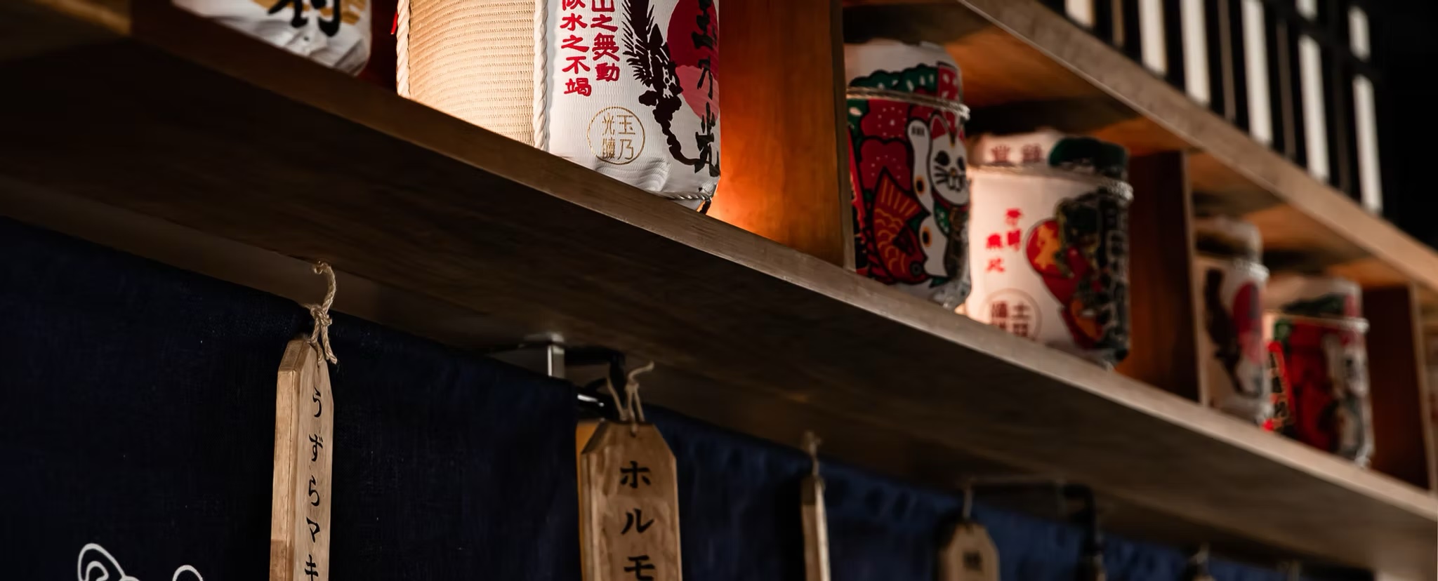 Sake barrels and lanterns lining a shelf above an indigo noren curtain